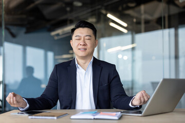 Asian young male office worker resting at his desk, sitting in lotus position and meditating with eyes closed