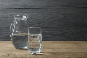 Glass of water with jug at wooden table, closeup. Drinking clear water in the morning for health. Space for text 