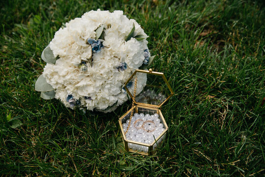 A bouquet of white roses lies on the green grass next to a golden box for wedding rings placed on the bottom strewn with pearls