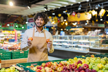 A smiling grocery store worker holds apples, standing in front of a display of fresh produce at a supermarket.