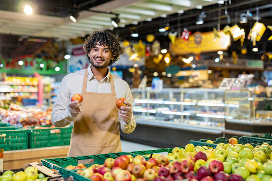 A smiling grocery store worker holds apples, standing in front of a display of fresh produce at a supermarket.