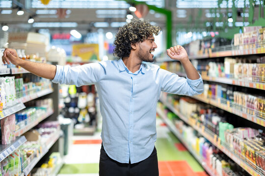 A cheerful man showing excitement while dancing in a supermarket with brightly lit aisles.