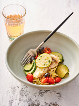 A bowl filled with spaghetti topped with freshly cooked cod, sliced zucchini, and vibrant cherry tomatoes. A glass of light beverage complements the meal, making it ideal for a casual lunch
