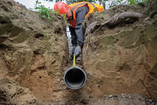 Construction worker in safety gear measuring and aligning a plastic drainage pipe in a trench during septic system installation at a private house, ensuring proper slope and connection for local sewag