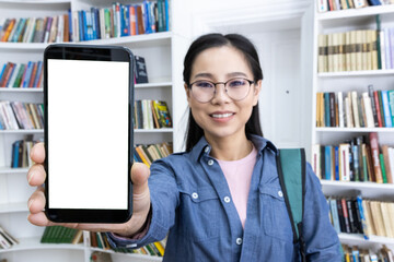 Smiling woman wearing glasses holding a phone with a blank screen while standing in a library with bookshelves in the background. Suitable for concepts related to technology