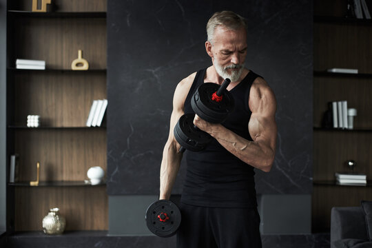 Middle aged Caucasian man exercising with dumbbells, showing muscular arms and focused expression, standing in modern interior, demonstrating strength and fitness lifestyle