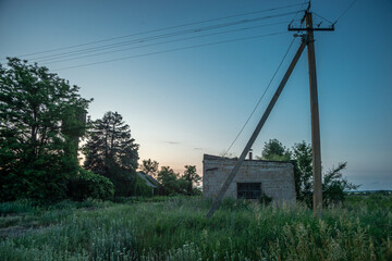A horizontal view of an abandoned brick building and a power line pole surrounded by overgrown nature and trees in the peaceful light of a summer evening, with a blue and pink gradient sky.