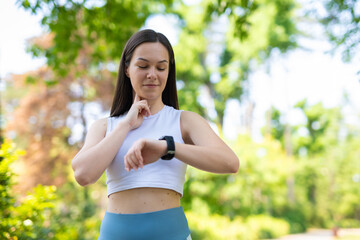 Fototapeta premium Young woman measuring pulse and checking smartwatch during outdoor workout in the park. 