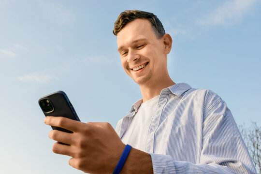 Smiling young man looking at smartphone outdoors on a sunny day, wearing casual shirt and enjoying good news against a clear blue sky