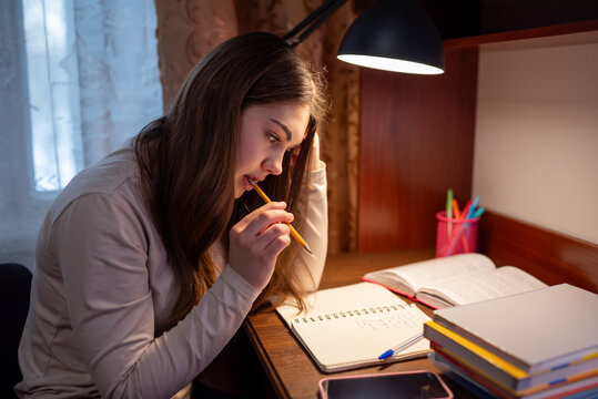 Student focused on completing her homework. A young woman sits at a desk with a pencil in her mouth, deep in thought and concentrated on her studies - Powered by Adobe
