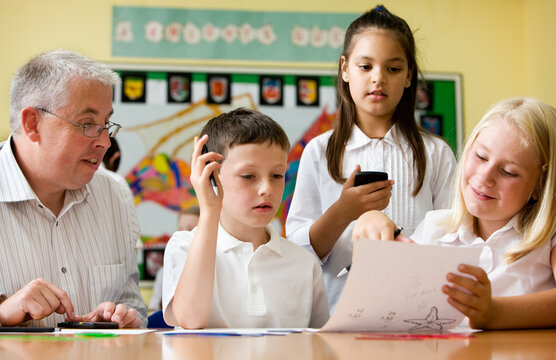 Primary school pupils using mobile phones for learning. A male teacher and a group of young students working on a task together at a classroom table. From an image series.