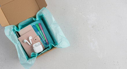 Top view of a student care package or employee welcome kit. Flat lay of a subscription box with stationery and wireless earbuds on a concrete background with ample copy space for text.