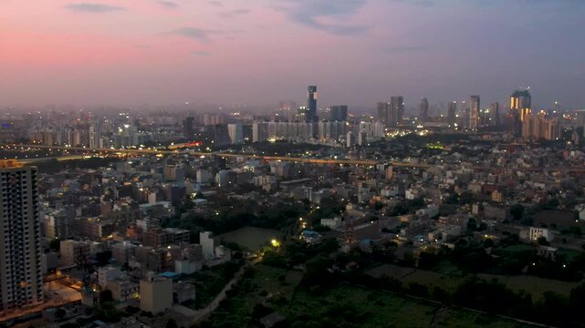 Aerial drone shot slow panning over gurgaon cityscape showing densely populated houses elevated roads, skyscrapers and water pools against the pink orange dusk sky
