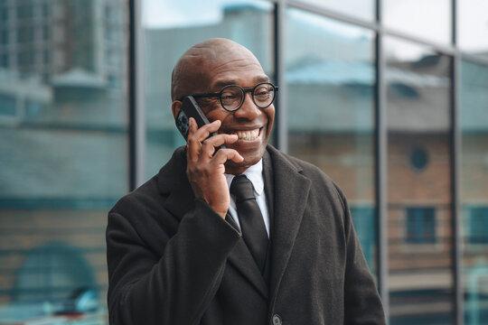 Man wearing glasses smiles while talking on a smartphone outside a modern building during daylight