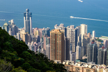 A panoramic view of Hong Kong dense cityscape, showcasing towering skyscrapers and a lush green hillside. The blue sky with a single fluffy cloud provides a picturesque backdrop.