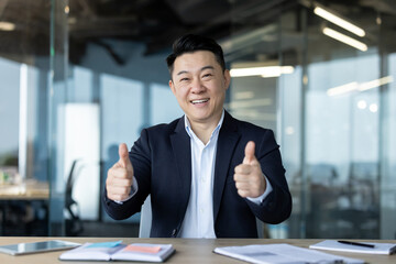 Portrait of a young Asian businessman sitting at his desk in the office and showing a super victory gesture to the camera
