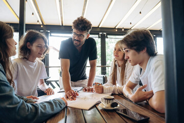 Guy in glasses is standing and explaining project plan. Group of young friends are in the cafe restaurant