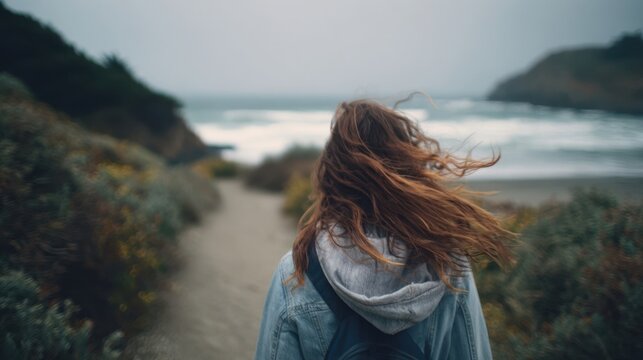Young woman tourist walking along a winding path toward the beach on a cloudy, windy day, with her hair dancing in the breeze, embracing the spirit of adventure and exploration