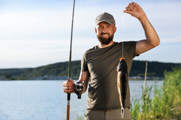 Smiling fisherman holding rod and caught fish near lake at summer