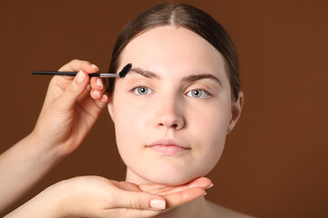 Fototapeta premium Cosmetologist brushing young woman's eyebrow with spoolie on brown background, closeup