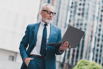 Confident senior businessman working on a laptop outdoors in an urban setting, standing near modern buildings.