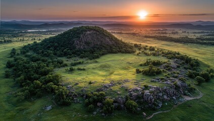 Sunrise over a grassy hilltop with rocky outcrops. Lush green vegetation surrounds a prominent, rounded hill with exposed rock formations.  A vibrant sunset paints the sky