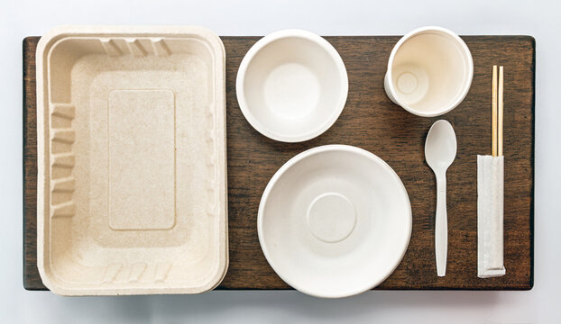 Eco friendly disposable tableware set includes tray, bowls, cup, spoon, and chopsticks on wooden surface, promoting sustainability