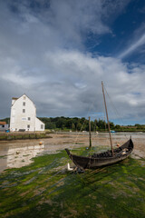 Old wooden boat with mast sitting on mud. Old tide mill behind. View of the quay at low tide in the River Deben at the historic town of Woodbridge in Suffolk, UK.