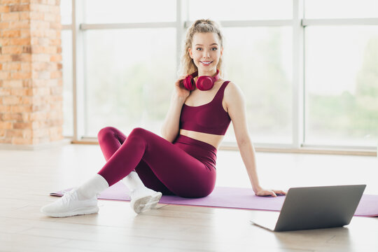 Sporty woman in activewear resting on mat with headphones and laptop near a bright window