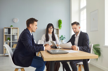 Three colleagues in formal attire sit at a wooden table in a bright modern office, reviewing documents and laptops. Focused teamwork drives discussion and planning. Modern space supports meeting.
