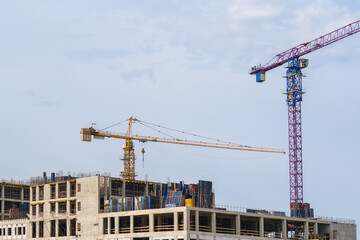 Two tall construction cranes overlooking the concrete structures of a building site under a clear sky. Real estate development.