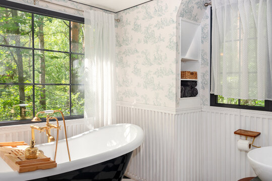Cozy bathroom with luxurious clawfoot bathtub, golden fixtures set near large windows overlooking a tranquil forest. Ideal for relaxation or spa-themed concepts, emphasizing comfort and serenity.