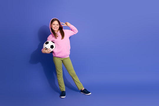 Happy schoolgirl holding a soccer ball, posing in a casual pink hoodie and green pants against a vibrant purple background.