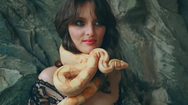fashion model girl posing with yellow snake hypnotizes wth eyes, sexy woman hypnosis. Studio background gray stone. Black long dress. Vampire gothic lady touch ancient albino boa constrictor animal