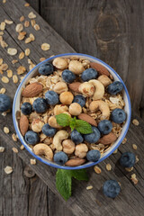Granola with nuts and fresh blueberries in glass bowl over old wood background.