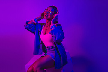 Obraz premium Young woman enjoying music with headphones in vibrant neon lighting, wearing stylish denim outfit, dancing joyfully