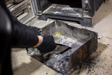 A man in black gloves cleans a metal ash pan with a scraper near a wood stove