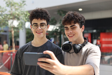 Two cheerful teenage twin brothers standing together and smiling while looking at a smartphone during a video call or live stream indoors. Casual urban youth lifestyle , friendship and family bonding