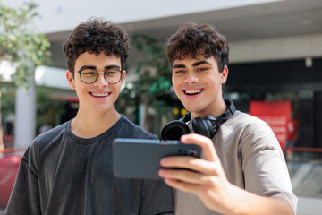 Two cheerful teenage twin brothers standing together and smiling while looking at a smartphone during a video call or live stream indoors. Casual urban youth lifestyle , friendship and family bonding