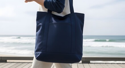A person carries a large navy blue tote bag while walking on a wooden boardwalk near a beach with ocean waves.