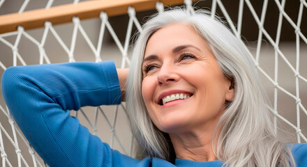 A happy, mature woman with long gray hair reclines in a hammock, enjoying a moment of relaxation and peace.
