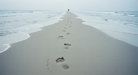A distant figure walking along a sandy beach leaving footprints in the sand under an overcast sky view