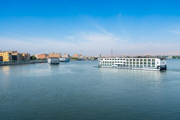 Fototapeta premium Multiple Nile River cruise ships navigate the calm waters, approaching the Esna Lock with the urban landscape of the city visible along the riverbank under a clear blue sky.