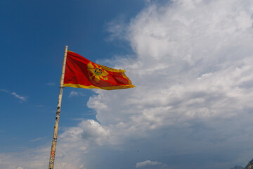 Montenegrin national flag waving on tall pole against dramatic cloudy sky with mountain peak visible on horizon, symbol of Montenegro independence and pride