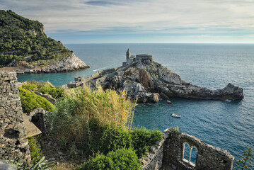  A view of the Church of St. Peter, also known as Loggia Romanica, perched on a dramatic rocky outcrop overlooking the sea, with ancient stone ruins and green cliffs in the foreground.