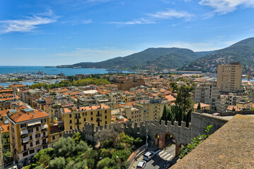 A panoramic view of the city of La Spezia and its harbor, seen from a castle, showing the densely packed buildings and the busy port surrounded by green hills under a partly cloudy sky