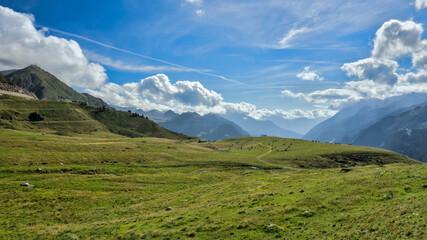 Switzerland Alps, Saint Gotthard: A view of a green, grassy mountain slope with a dirt path, and a wide mountain landscape in the background with cloudy sky.