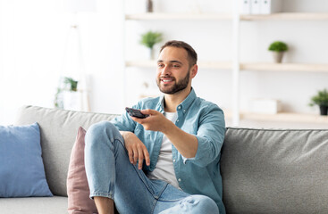 Weekend leisure activities. Millennial Caucasian man with remote control switching channels on TV at home. Happy young guy watching interesting program on television indoors