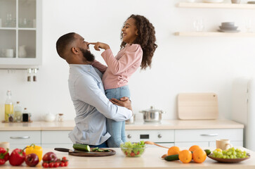 Happy Loving Family. Cute Curly Black Girl And Her Dad Fooling Together While Cooking In Kitchen, Preparing Healthy Salad, Man Holding And Hugging Daughter Who Playfully Touching Father's Nose