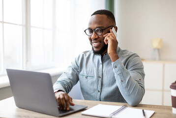 Fototapeta premium Happy black businessman talking on cellphone having phone conversation sitting at workplace working on laptop computer in office. Man chatting on smartphone indoor. Mobile communication concept
